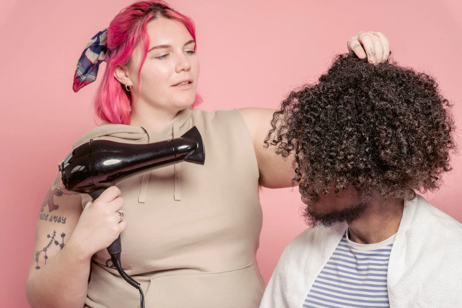 Hairdresser blow-drying a client's curly afro hair against a pink background